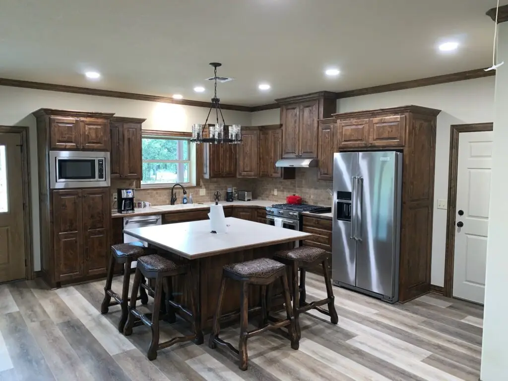 Shows the inside of a house, there is gray wood-like linoleum flooring, dark brown cabinets and a matching island. There are dark brown wooden barstools with worked leather seats.